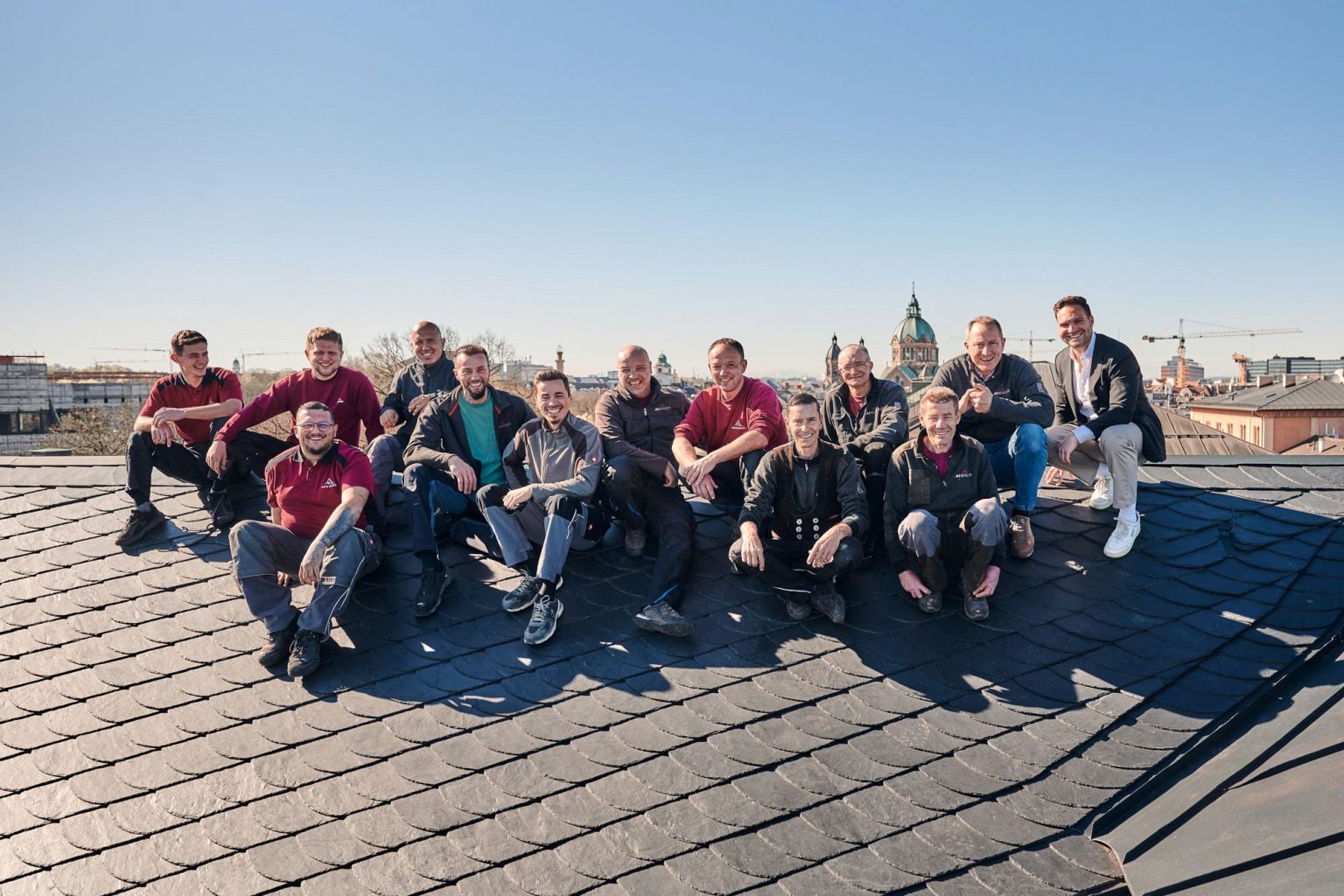 Gruppe von Handwerkern posiert auf einem Dach mit Dachziegeln, im Hintergrund München bei klarem Himmel.