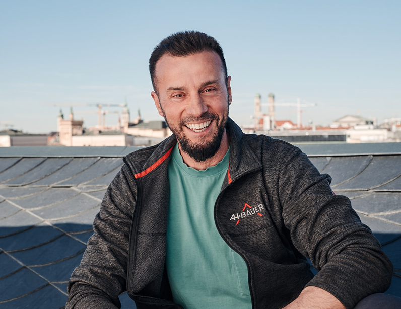 Lächelnder Mann mit Bart in Arbeitskleidung, vor einer Stadtlandschaft mit Baukränen im Hintergrund.