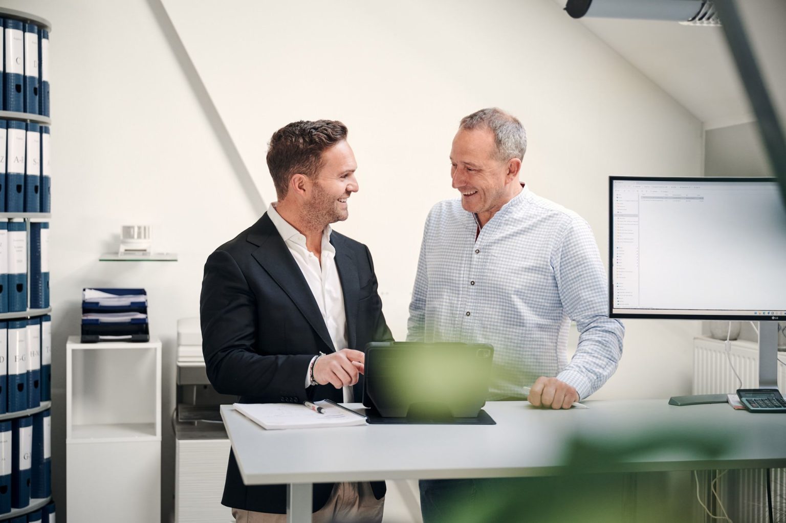 Geschäftsführer und Betriebsleiter diskutieren in einem modernen Büro lächelnd über ein Tablet, umgeben von Aktenordnern und einem Computer.