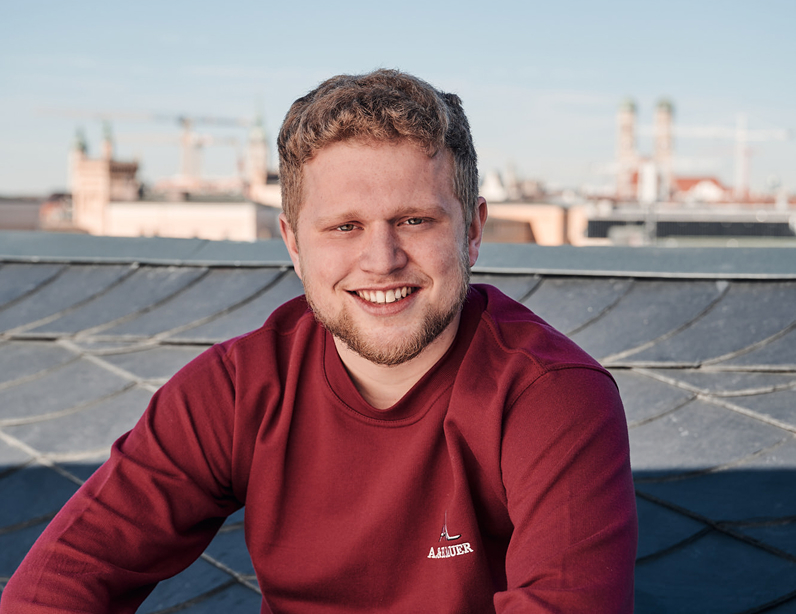 Lächelnder junger Mann mit lockigem Haar sitzt auf einem Dach, im Hintergrund eine Stadtlandschaft mit Baukränen.