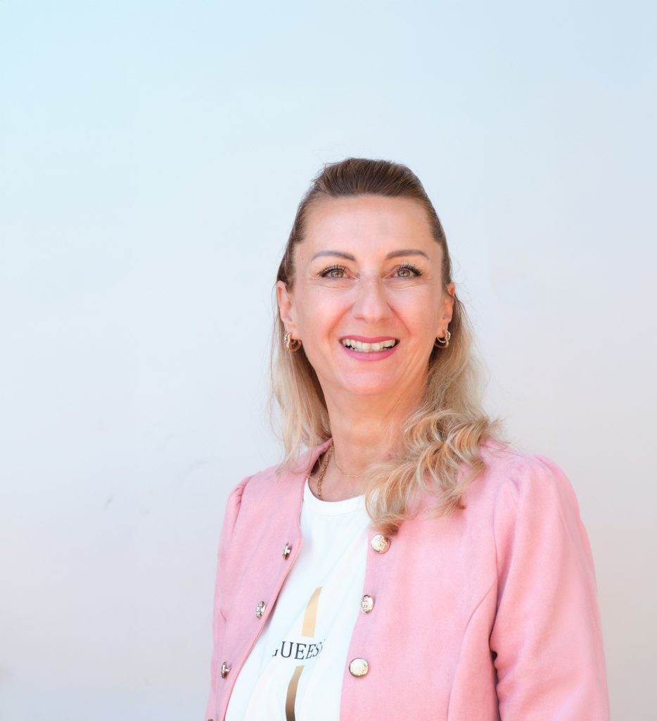 Michaela - Assistenz der Geschäftsleitung. Miit blonden Haaren und rosa Jacke lächelt sie vor einem hellen Hintergrund und strahlt Freundlichkeit aus.