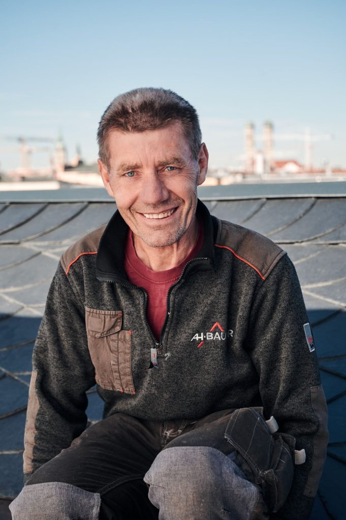 Handwerker Johann sitzt auf dem Dach, lächelt freundlich in die Kamera, im Hintergrund eine Stadtlandschaft bei klarem Himmel.