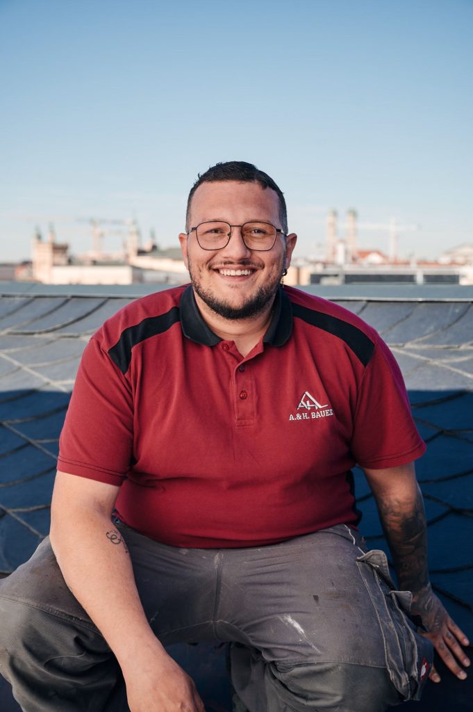 Handwerker Antonio sitzt in rot-schwarzem Polo-Shirt auf dem Dach, lächelt vor urbaner Kulisse mit Baukränen im Hintergrund.