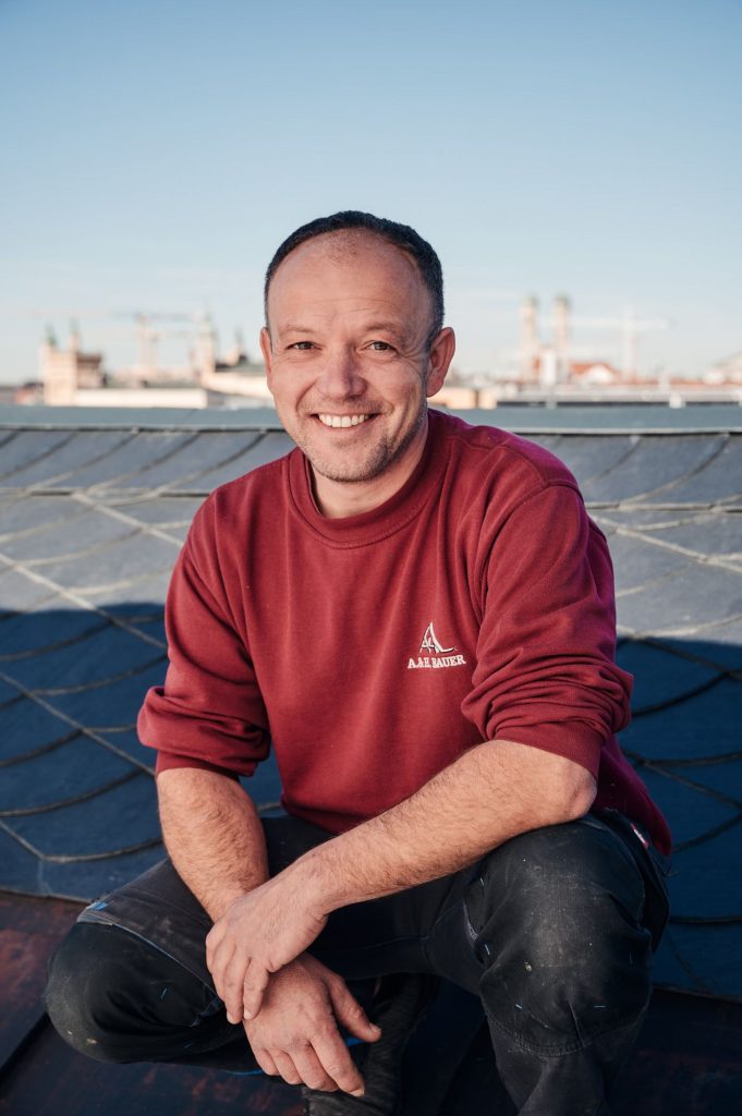 Handwerker Tobias auf dem Dach, lächelnd, in rotem Pullover, mit Baustellenhintergrund und klarem Himmel.