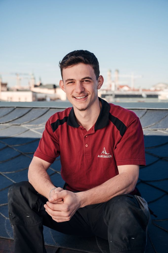 Handwerker Ali in rotem Poloshirt sitzt auf einem Dach, mit Stadtlandschaft im Hintergrund und lächelt freundlich.