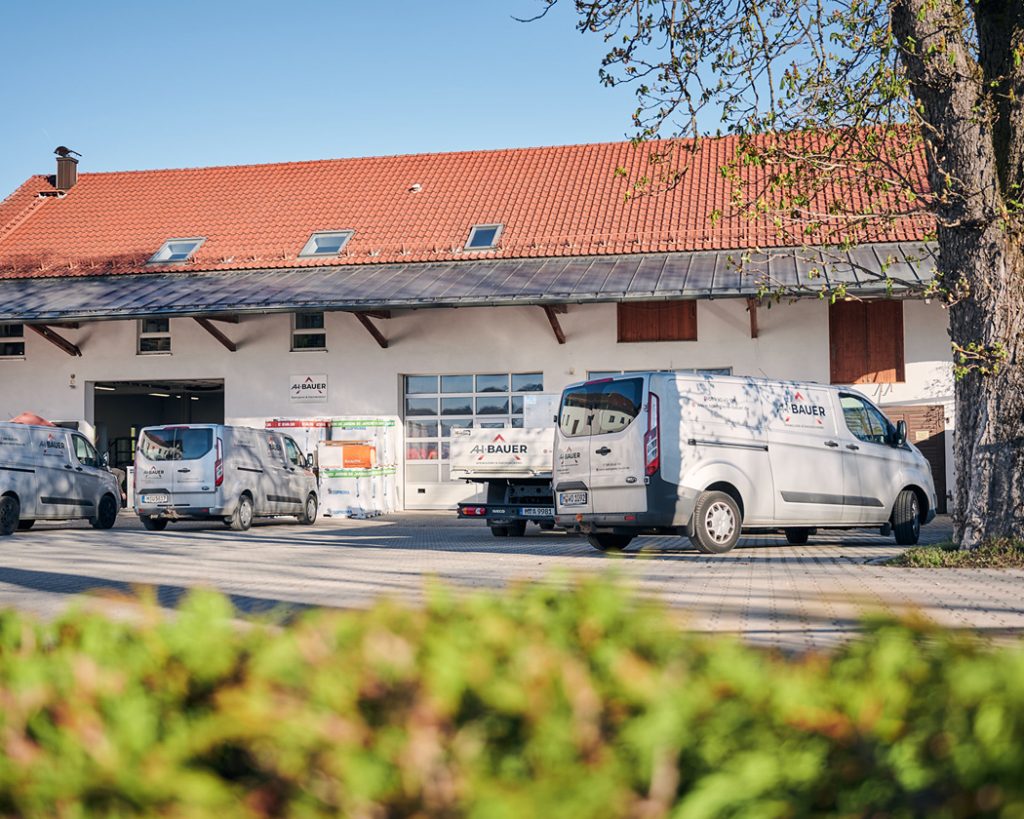 Bauer Firmengebäude mit mehreren Lieferwagen auf dem Parkplatz unter blauem Himmel und frischem Grün im Vordergrund.