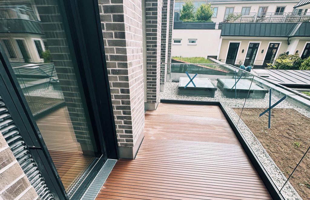 Moderne Terrasse mit Holzbelag, Glasgeländer und Blick auf bepflanzte Flächen in einem urbanen Wohngebiet.