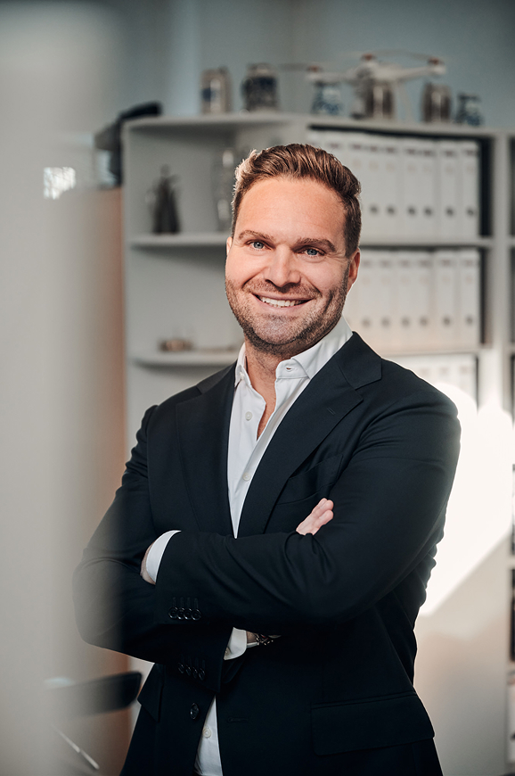 Professioneller Mann in Anzug lächelt selbstbewusst vor einem modernen Büro mit Aktenregalen im Hintergrund.