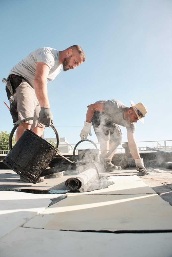 Zwei Handwerker tragen Dachmaterialien auf, während sie auf einem sonnigen Dach arbeiten und dampfende Materialien verwenden.