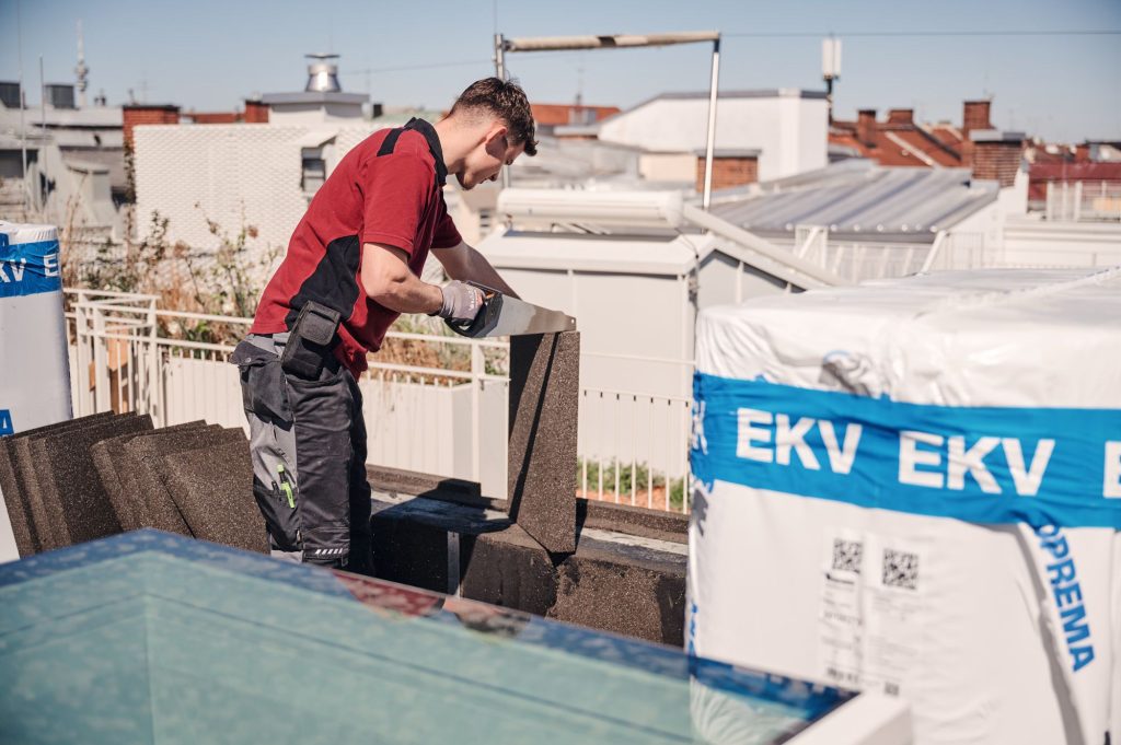 Arbeiter auf dem Dach schneidet Dämmmaterial, umgeben von Baustellenmaterial und moderner Architektur.