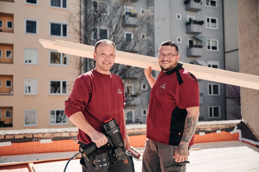 Zwei Handwerker auf einer Baustelle mit Holzlatten und Werkzeug. Ein Wohngebäude im Hintergrund.