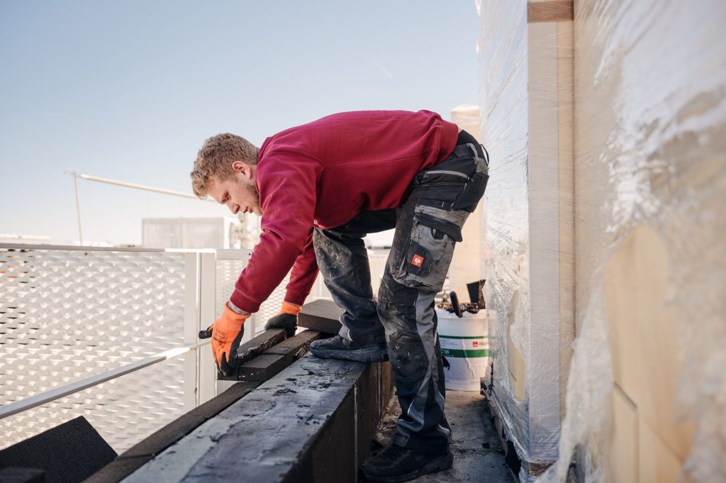 Handwerker verlegt Dachmaterial auf einer Baustelle, trägt Arbeitshandschuhe und konzentriert sich auf seine Aufgabe.