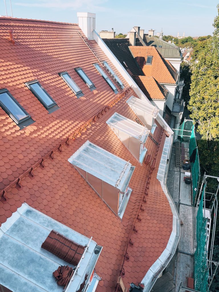Dachansicht mit roten Biberschwänzen, Dachfenstern und Baugerüst, umgeben von städtischen Gebäuden und Bäumen.