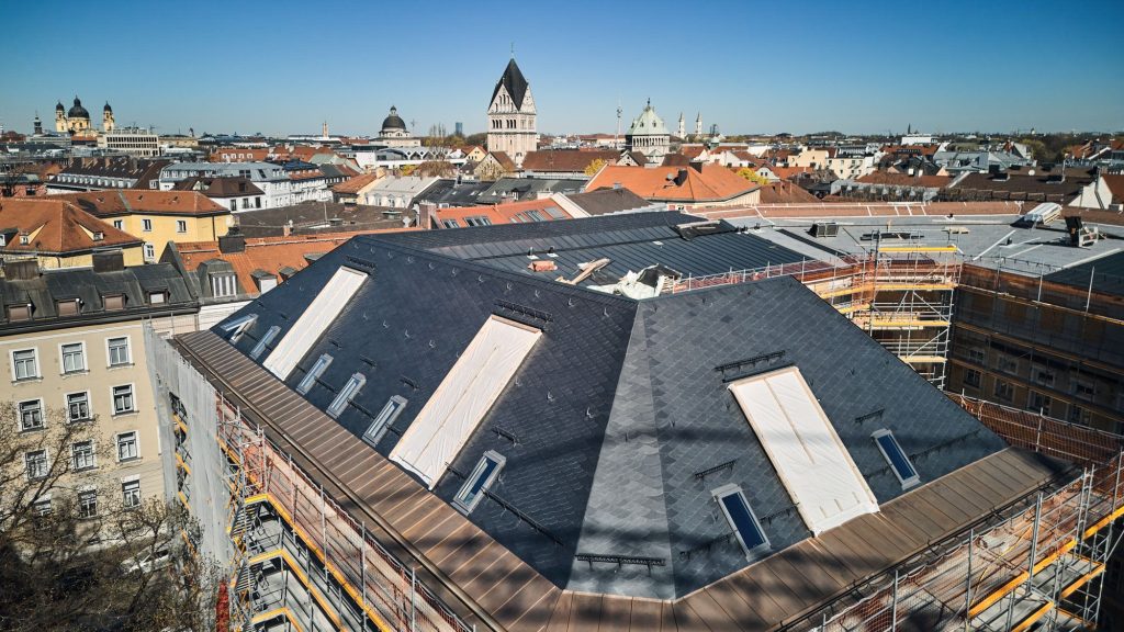 Blick auf eine große Baustelle mit Schieferdach und großen Dachfenstern. Umgeben von historischen Gebäuden in einer Stadt unter blauem Himmel.