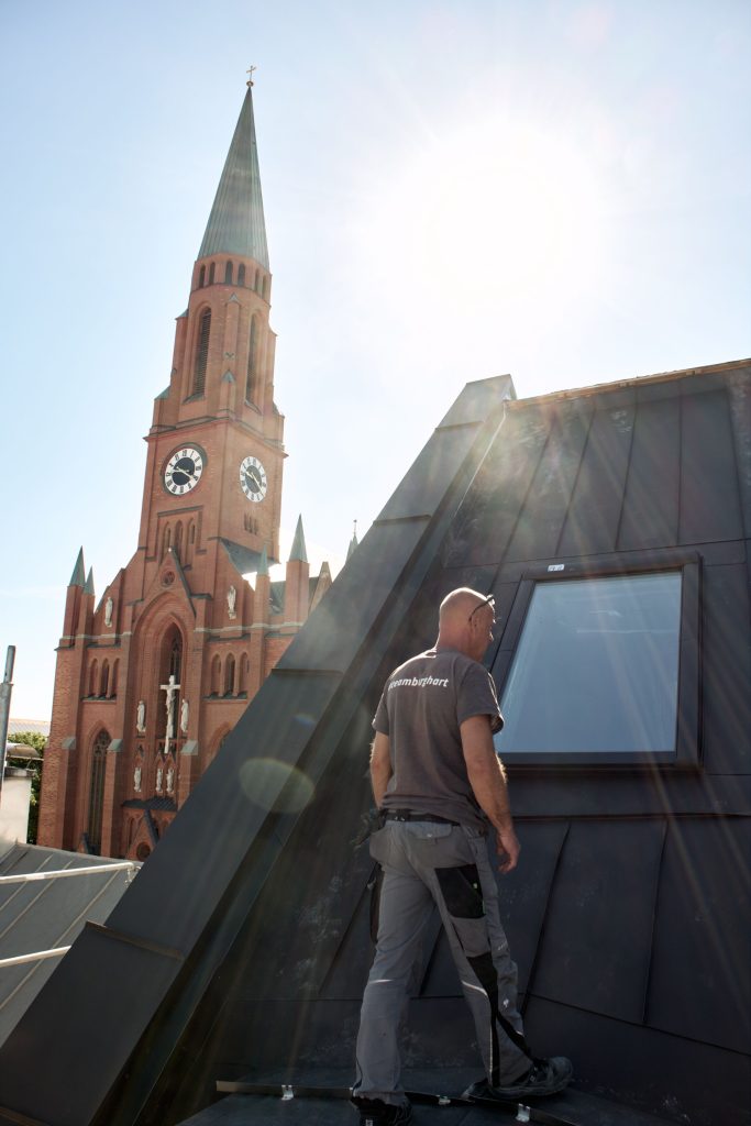 Dachdecker arbeitet an einem modernen Dach mit Blick auf die Kirche im Hintergrund, strahlende Sonne am Himmel.