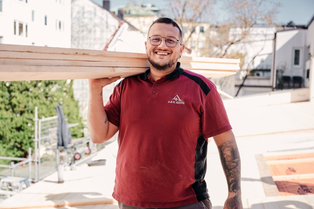 Handwerker trägt Holzplatten auf der Baustelle, lächelt und zeigt Einsatz.