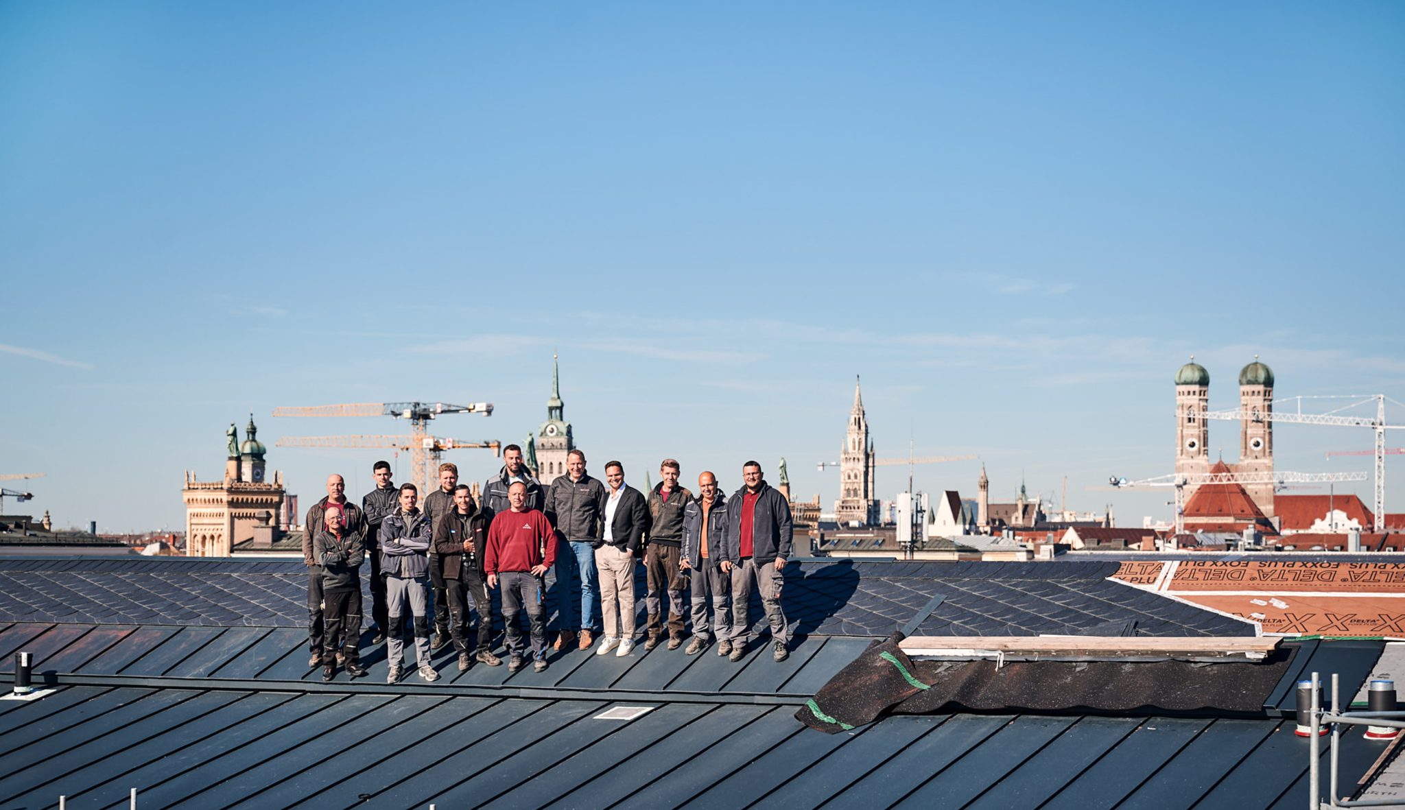 Gruppe von Handwerkern steht auf einem Dach mit Dachziegeln, im Hintergrund die Skyline von München.