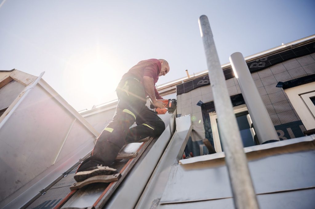 Handwerker auf einer Leiter bei der Montage von Blechelementen an einem Gebäude unter klarem Himmel.