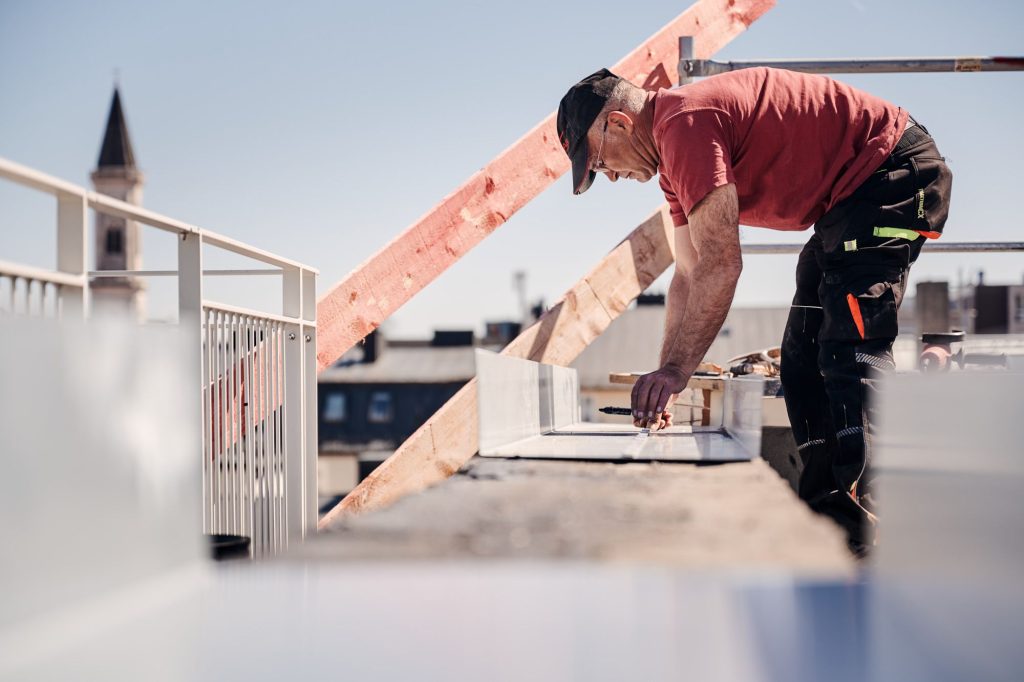 Handwerker arbeitet auf dem Dach und misst Materialien zur Renovierung, im Hintergrund ist ein Kirchturm sichtbar.