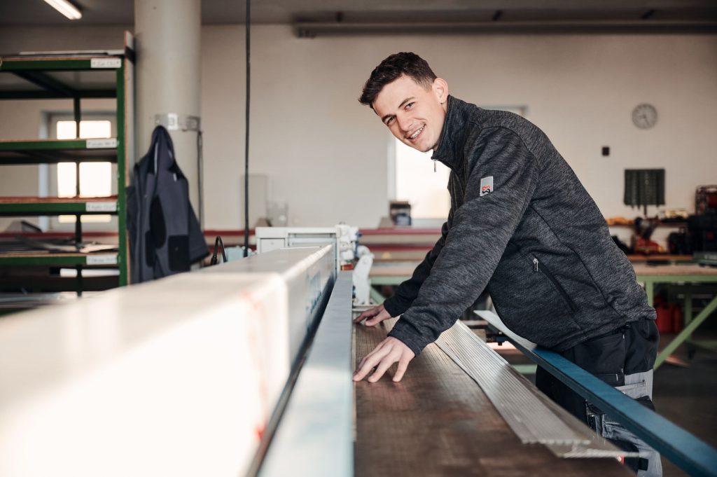 Junger Mann in Werkstatt, der an einer Maschine arbeitet und freundlich in die Kamera lächelt. Handwerkliche Tätigkeit und Präzision.