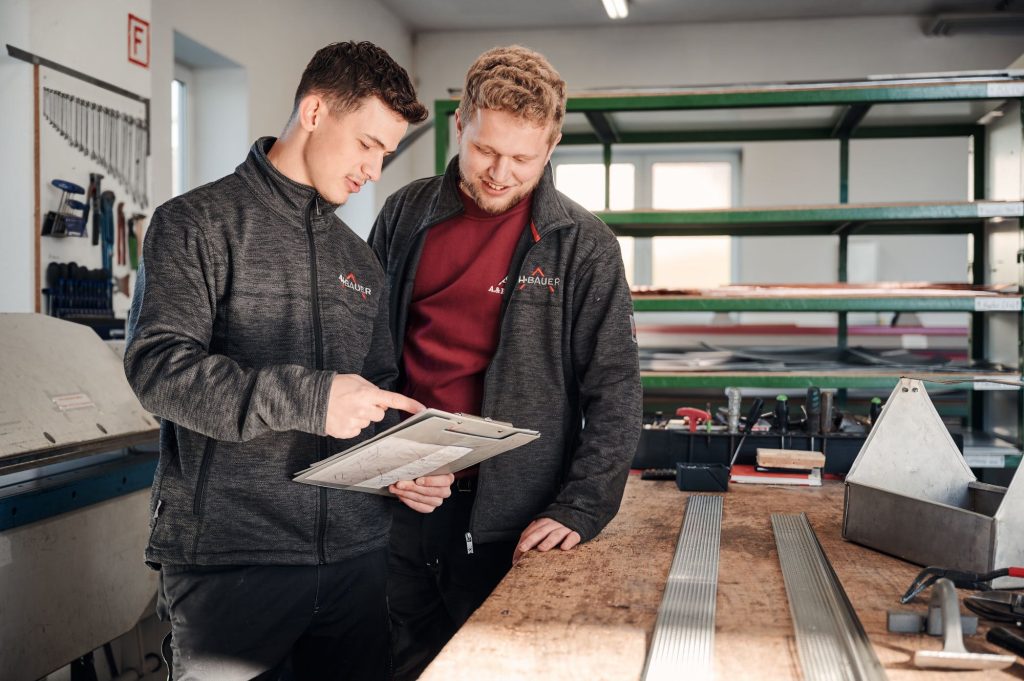 Zwei Männer in Arbeitskleidung besprechen Pläne in einer Werkstatt, umgeben von Werkzeugen und Materialien.
